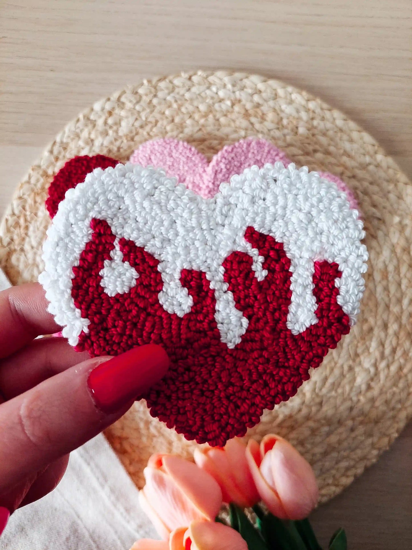 Handmade punch needle coaster shaped like a white heart with red flames detail, displayed on a woven mat with decorative flowers in the back.