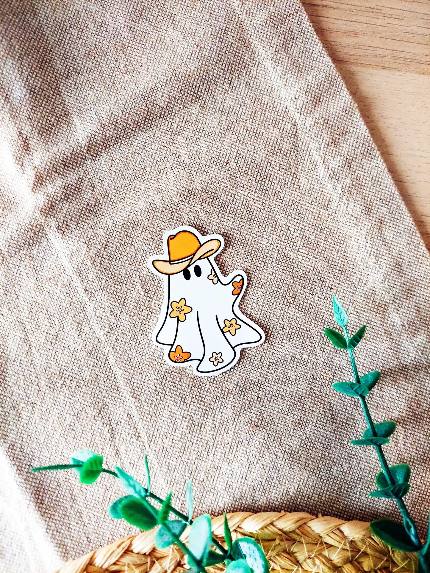 Vinyl sticker of a cute ghost wearing a cowboy hat with small orange flowers, photographed on a woven neutral background.