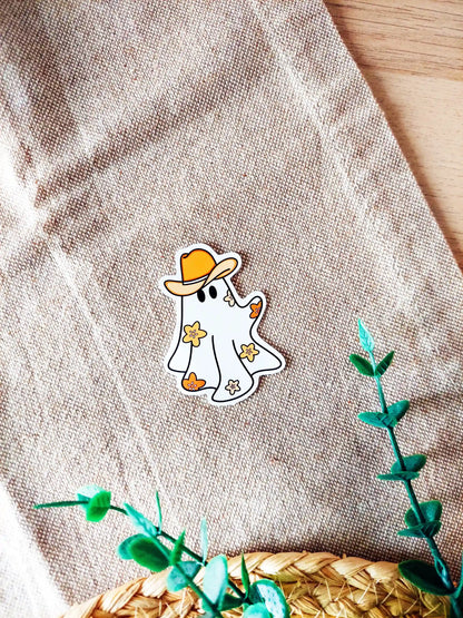 Vinyl sticker of a cute ghost wearing a cowboy hat with small orange flowers, photographed on a woven neutral background.