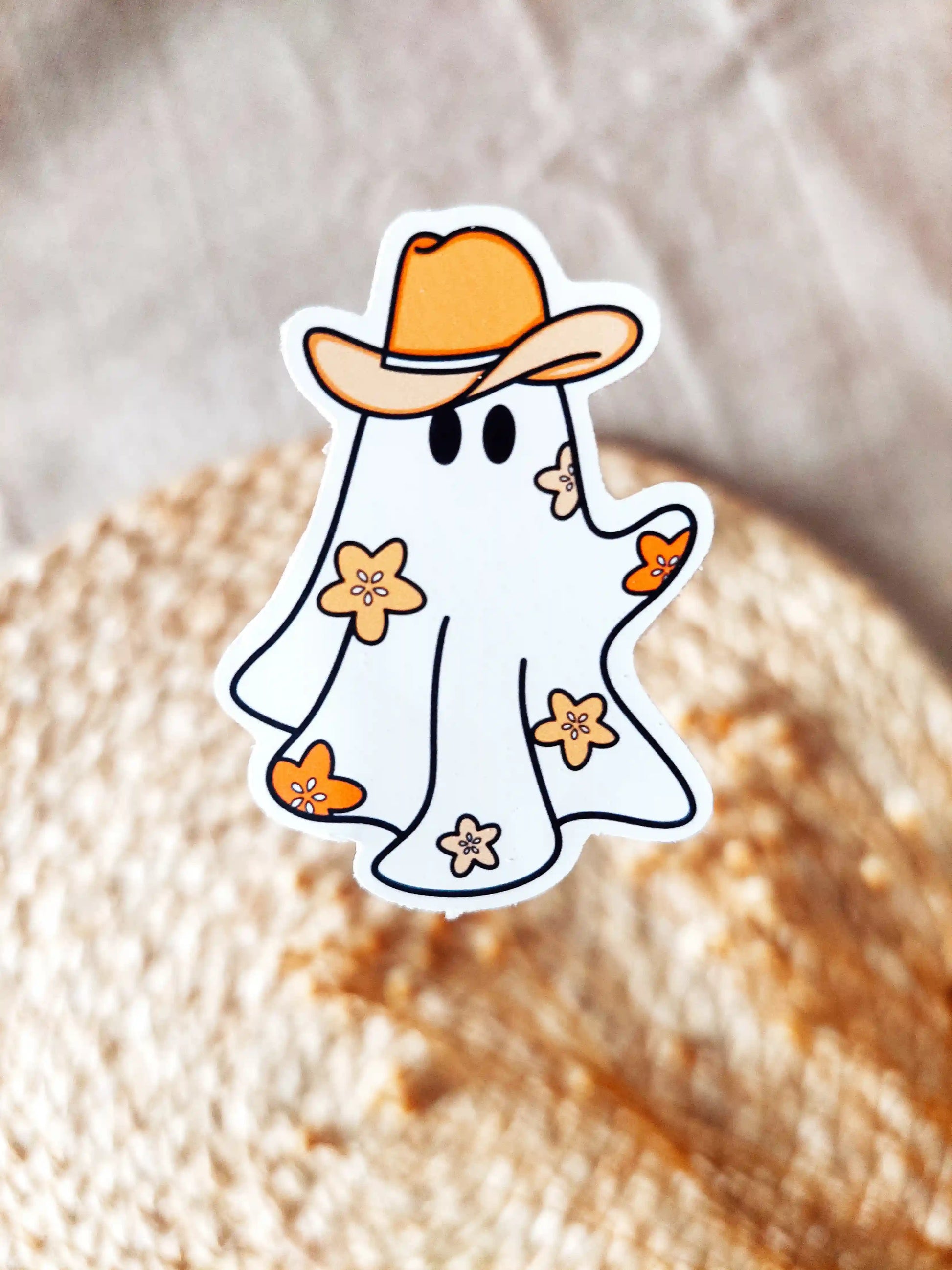 Vinyl sticker of a cute ghost wearing a cowboy hat with small orange flowers, photographed on a woven neutral background.