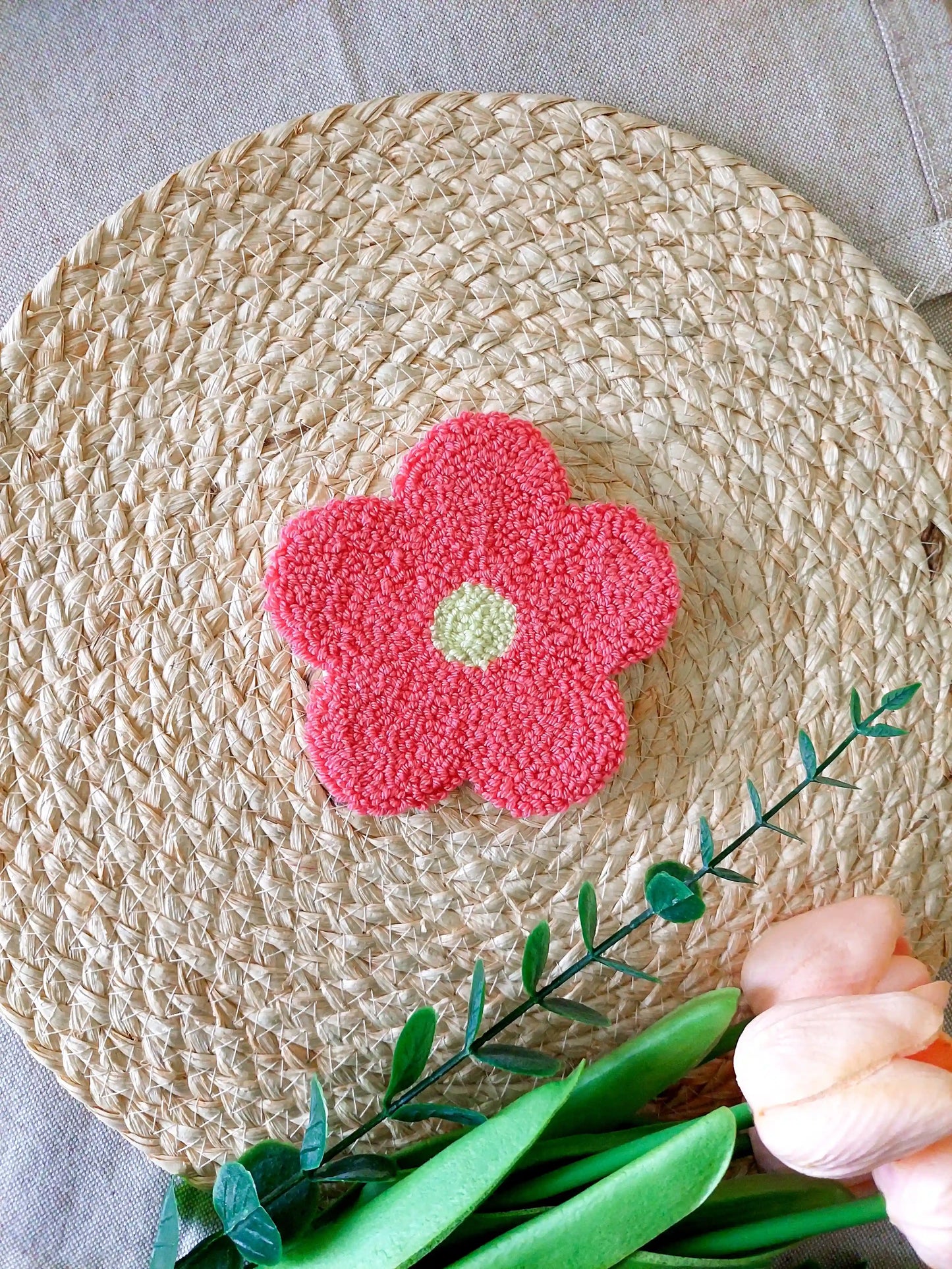 Handmade punch needle flower coaster with coral pink petals and a yllow center, photographed on a woven mat with decorative flowers in the back.
