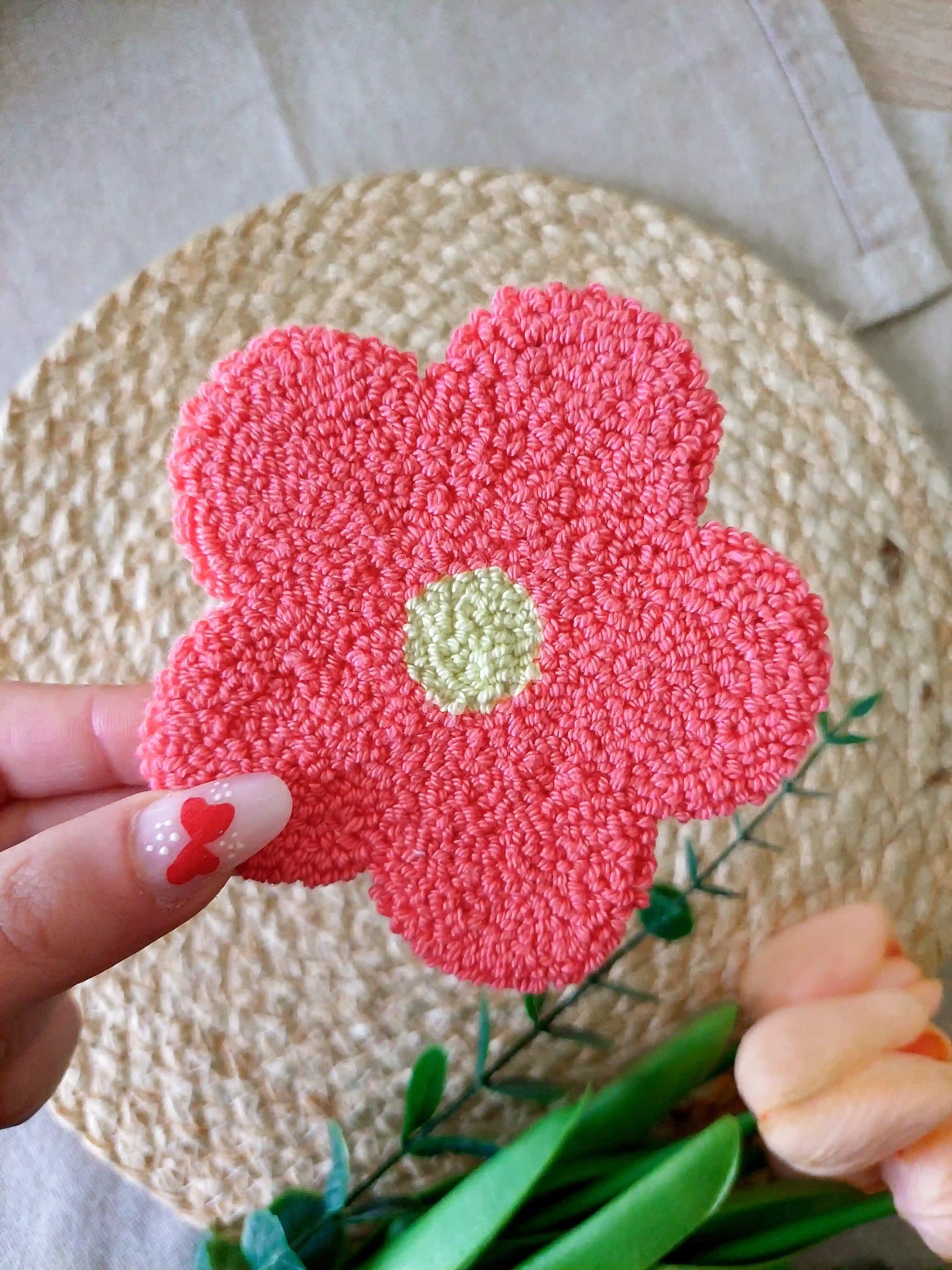 Handmade punch needle flower coaster with coral pink petals and a yllow center, photographed on a woven mat with decorative flowers in the back.