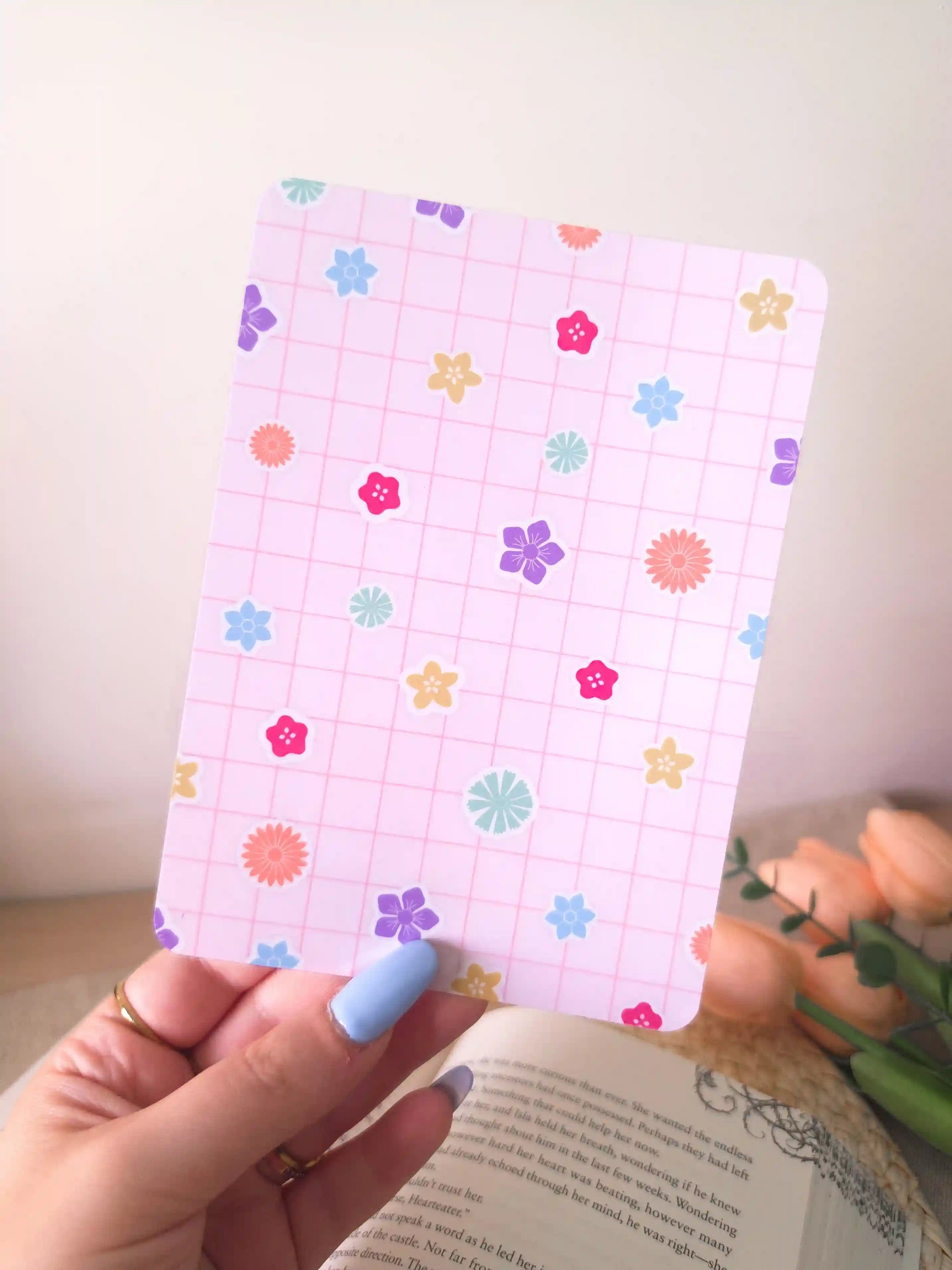 A hand holding an e-reader insert with a soft lavender grid background scattered with colorful flower illustrations in pink, blue, orange, purple, green, and yellow, placed on an open book resting on a woven rattan mat. 