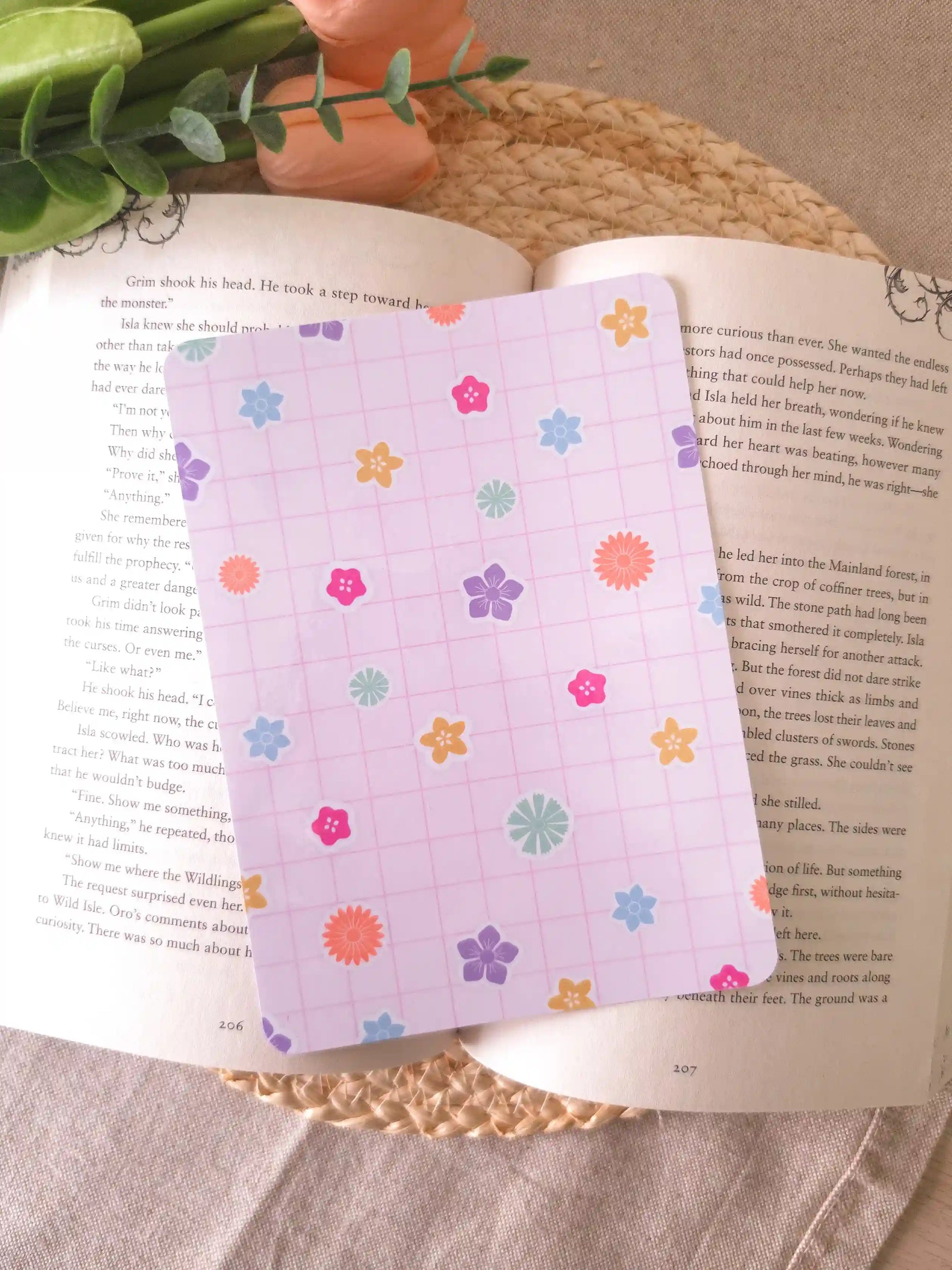 An e-reader insert with a soft lavender grid background scattered with colorful flower illustrations in pink, blue, orange, purple, green, and yellow, placed on an open book resting on a woven rattan mat. 