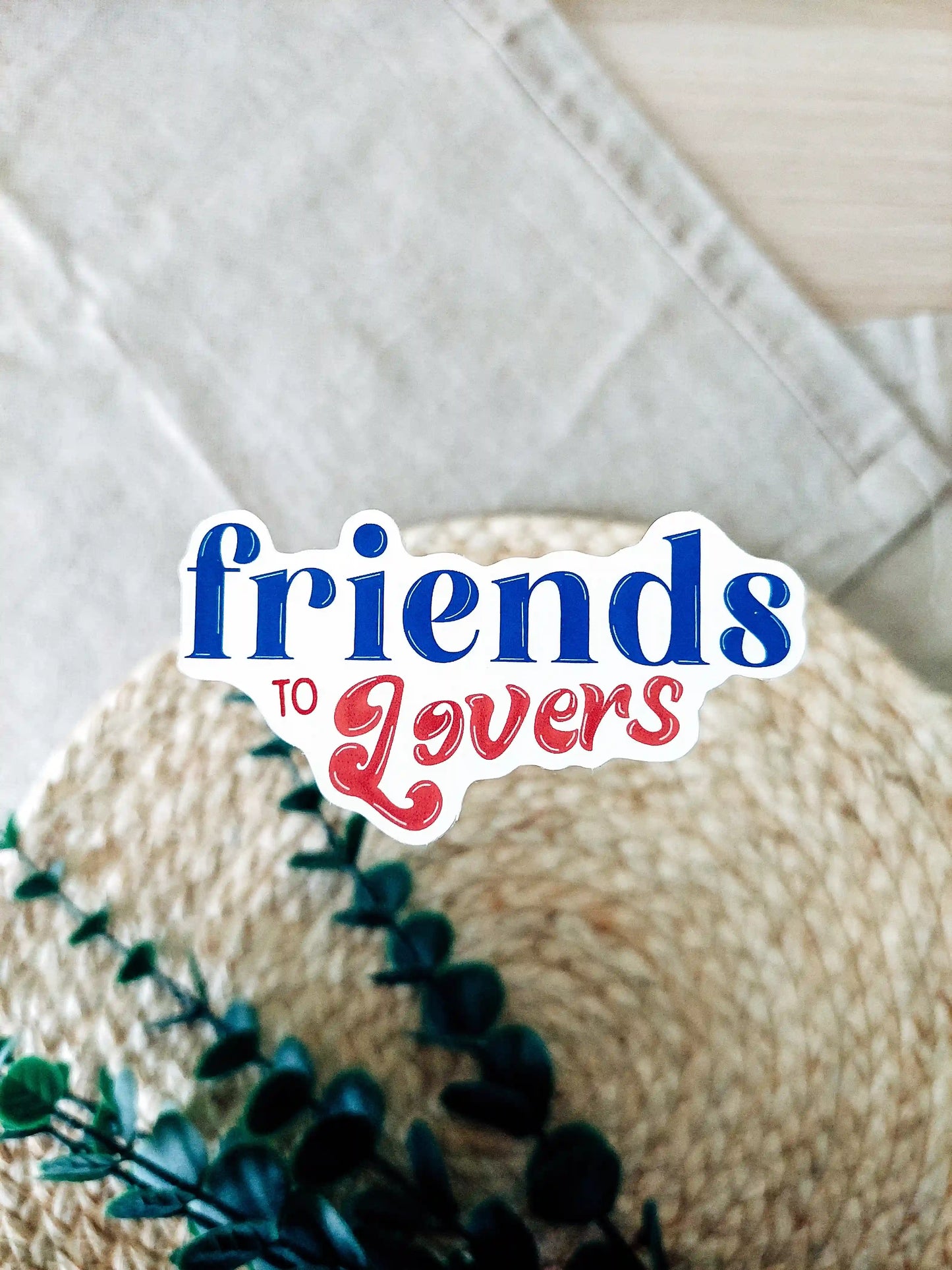 Vinyl sticker with the text "Friends to Lovers" in blue and red typography, photographed on neutral fabric background with woven mat and green foliage.