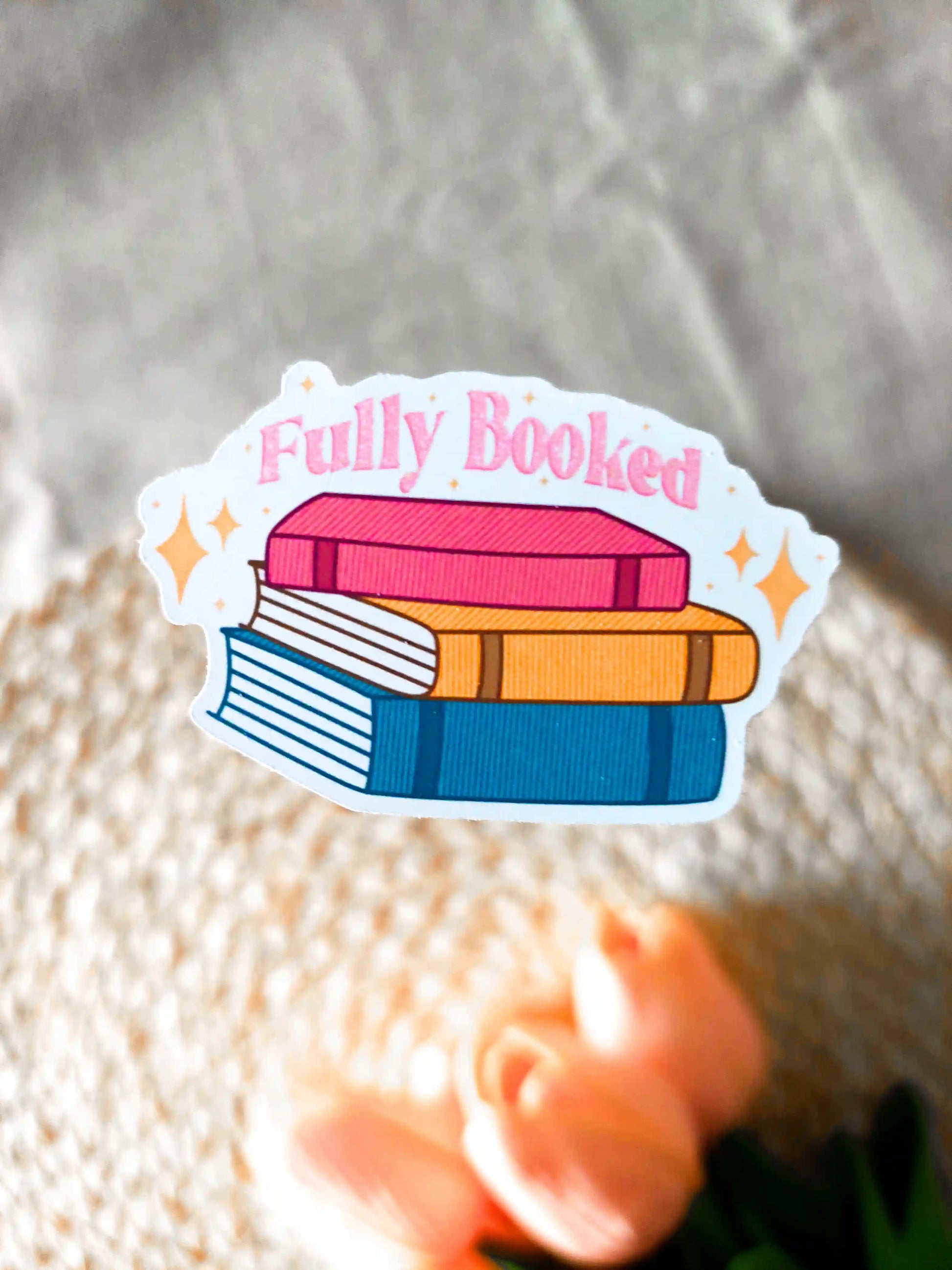 Vinyl sticker featuring a colorful stack of books with the phrase "Fully Booked", photographed on a woven neutral background.