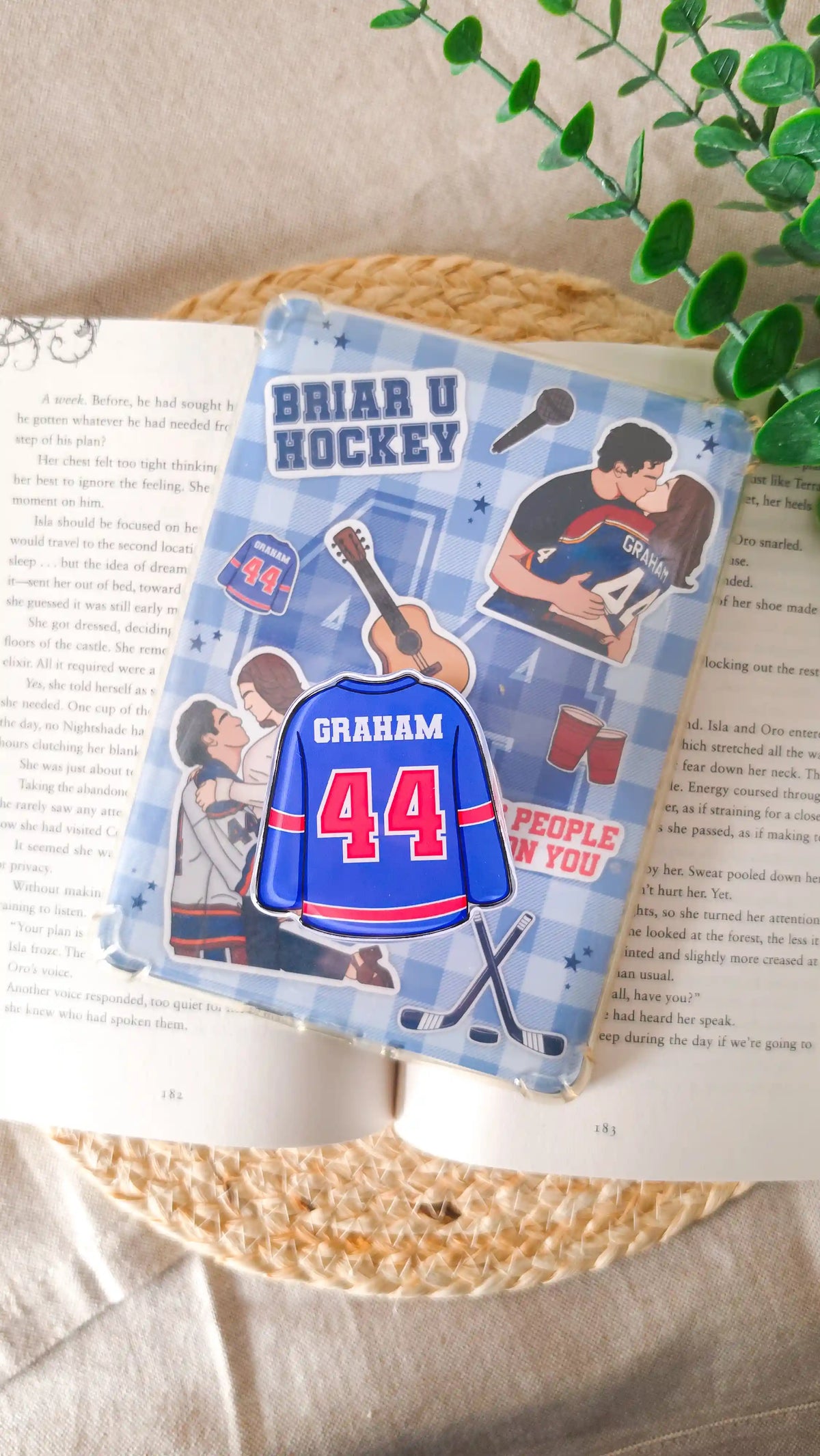 Blue hockey jersey magnetic grip with “Graham 44" attached to an e-reader with a blue checkered Off Campus insert showing illustrated couples, a guitar, red cups and crossed hockey sticks, lying on an open book on a woven rattan mat with green leaves in the corner.