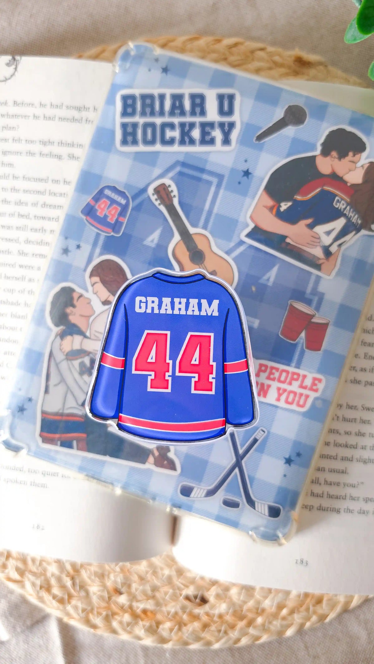 Blue hockey jersey magnetic grip with “Graham 44" attached to an e-reader with a blue checkered Off Campus insert showing illustrated couples, a guitar, red cups and crossed hockey sticks, lying on an open book on a woven rattan mat with green leaves in the corner.