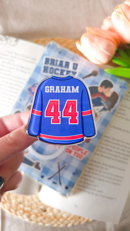 Hand holding a blue acrylic hockey jersey phone grip with “Graham 44”, in front of a blue checkered Off Campus e-reader insert. 