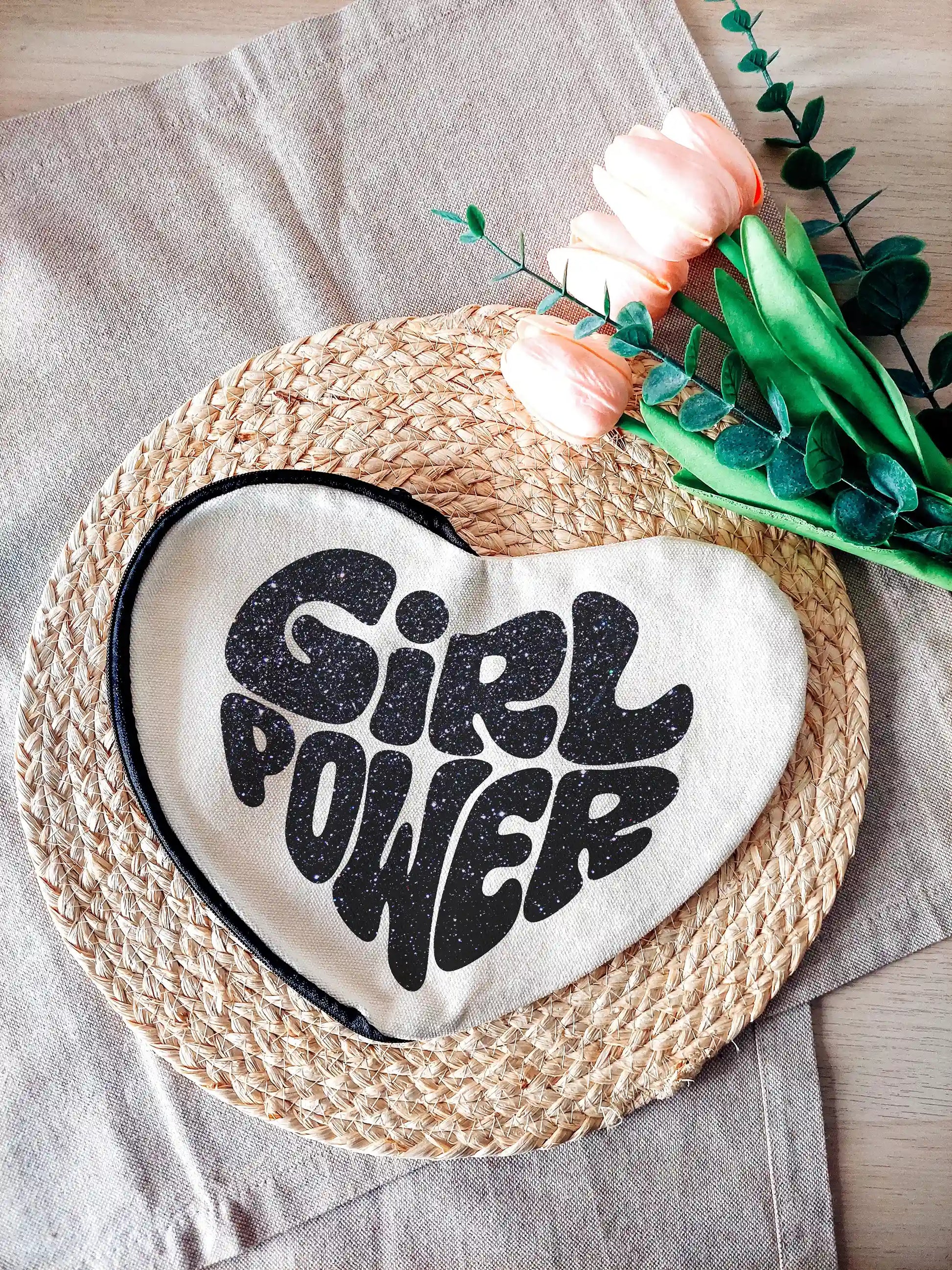 Heart-shaped fabric pouch with glittery black "Girl Power" text, photographed on a neutral background with decorative flowers on the right.