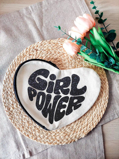 Heart-shaped fabric pouch with glittery black "Girl Power" text, photographed on a neutral background with decorative flowers on the right.