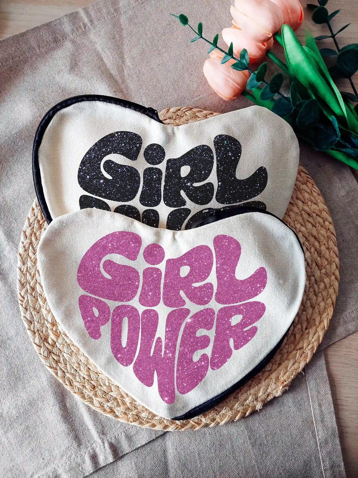Heart-shaped fabric pouches with glittery pink or black "Girl Power" text, photographed on a neutral background with decorative flowers on the right.