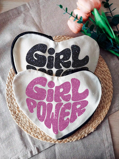 Heart-shaped fabric pouches with glittery pink or black "Girl Power" text, photographed on a neutral background with decorative flowers on the right.