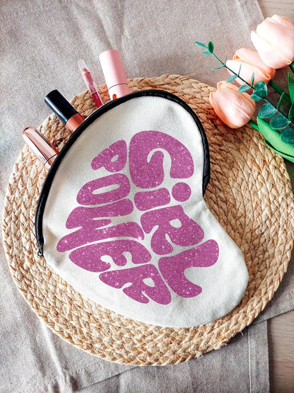 Heart-shaped fabric pouch with glittery pink  "Girl Power" text, partially unzipped and filled with makeup items, photographed on a neutral background with decorative flowers on the right.