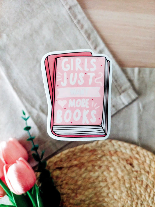 Vinyl sticker of a pink book with white text "Girls Just Want More Books", photographed on a neutral fabric background with tulips.