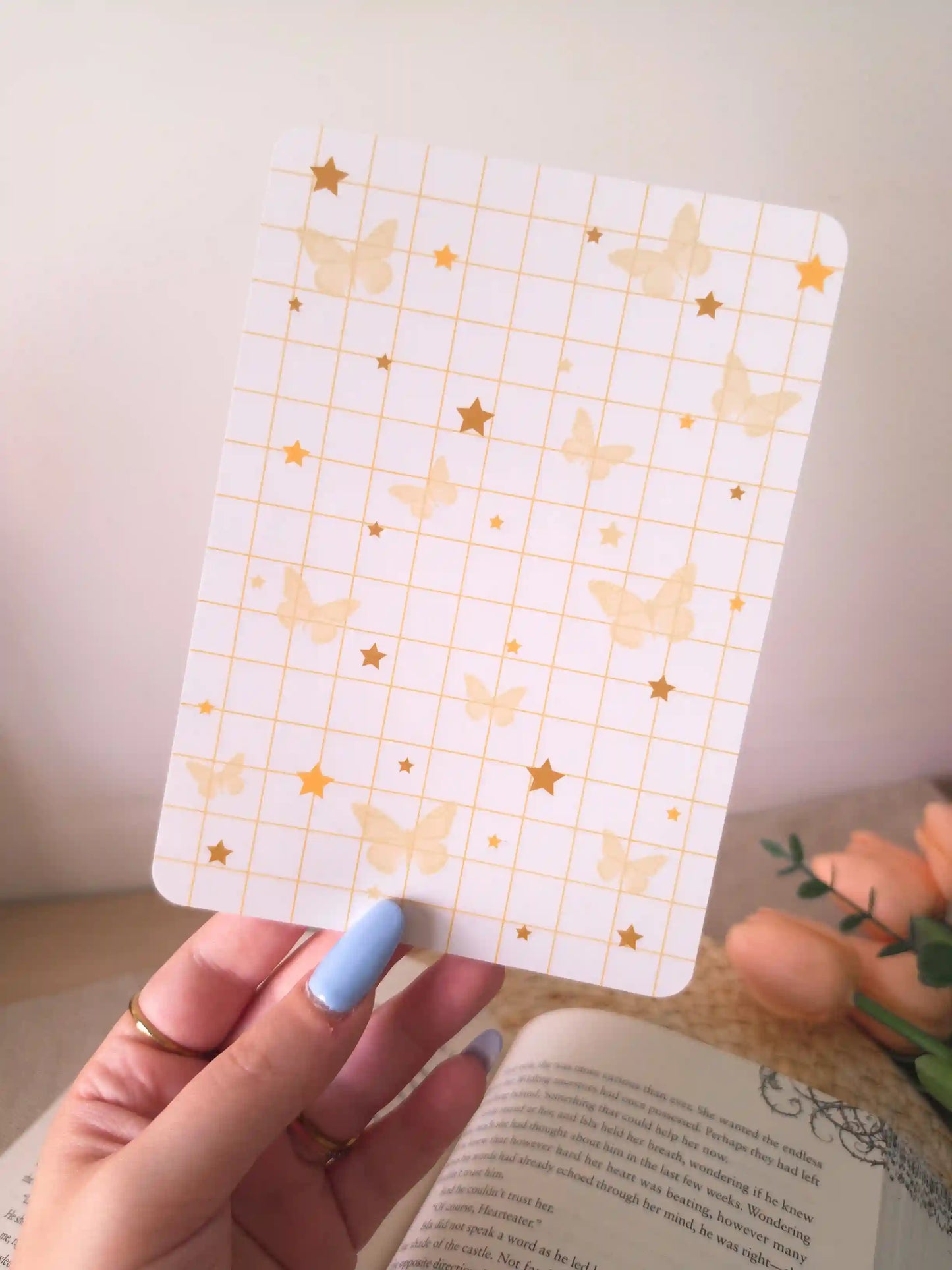 A hand holding an e-reader insert with a white grid background scattered with soft golden butterfly illustrations and gold star details in various sizes, placed on an open book resting on a woven rattan mat. 