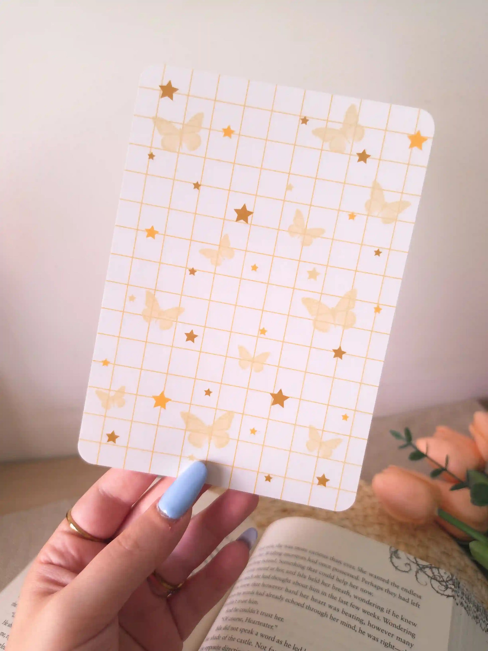 A hand holding an e-reader insert with a white grid background scattered with soft golden butterfly illustrations and gold star details in various sizes, placed on an open book resting on a woven rattan mat. 