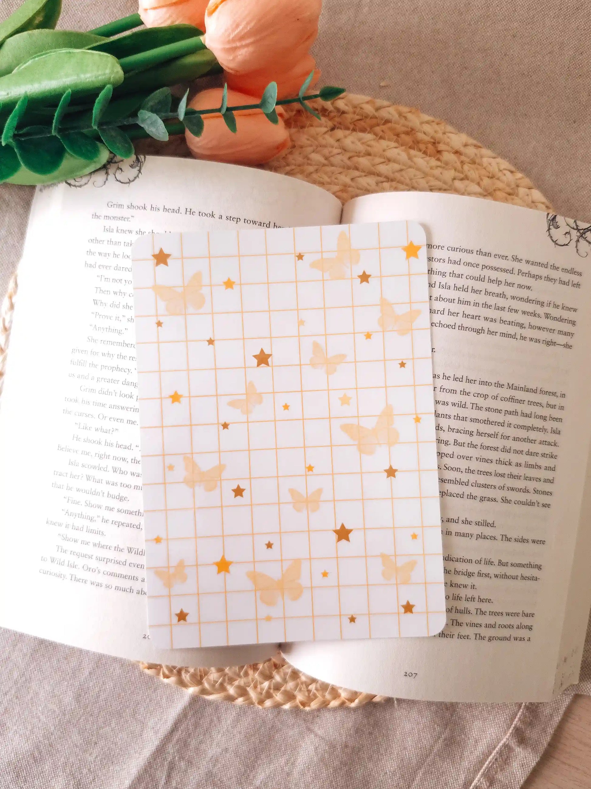 An e-reader insert with a white grid background scattered with soft golden butterfly illustrations and gold star details in various sizes, placed on an open book resting on a woven rattan mat. 