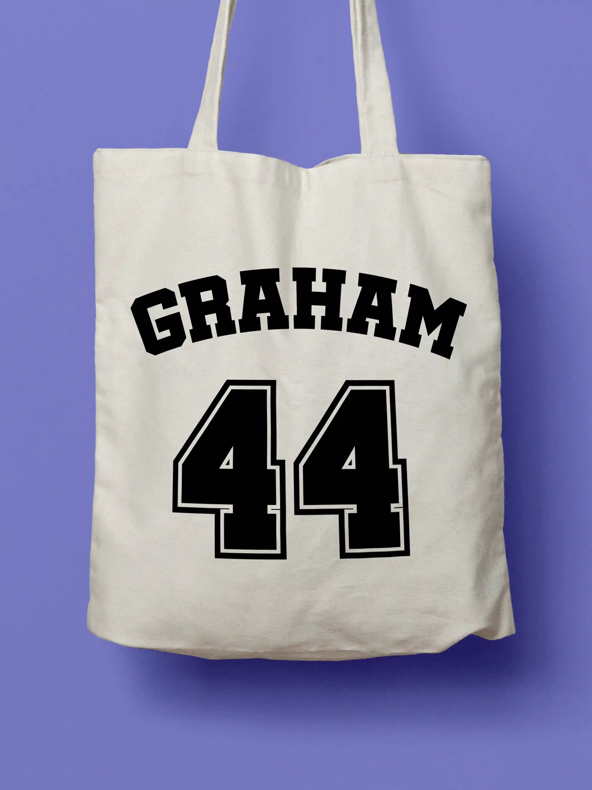 A white canvas tote bag featuring "Graham 44" in bold black collegiate-style lettering, photographed against a lavender background.