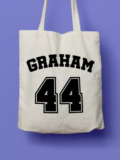 A white canvas tote bag featuring "Graham 44" in bold black collegiate-style lettering, photographed against a lavender background.
