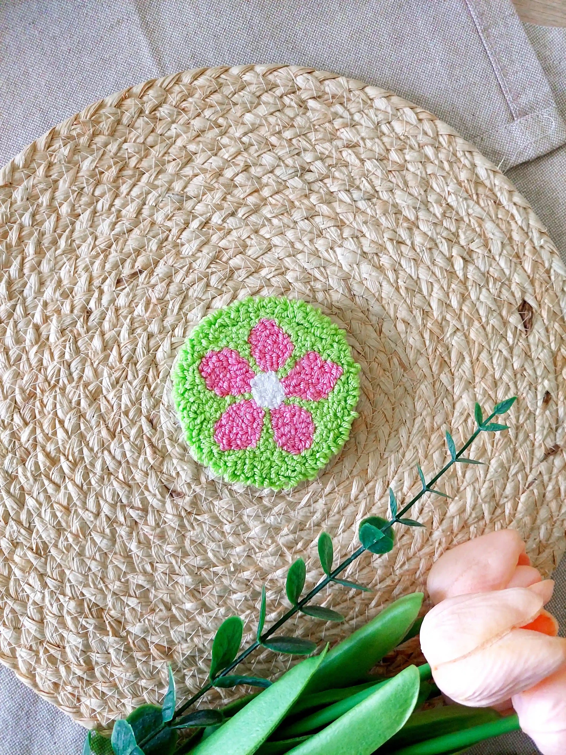 Handmade punch needle coaster featuring a bright green background with a pink flower, photographed on a woven mat with decorative flowers in the back.