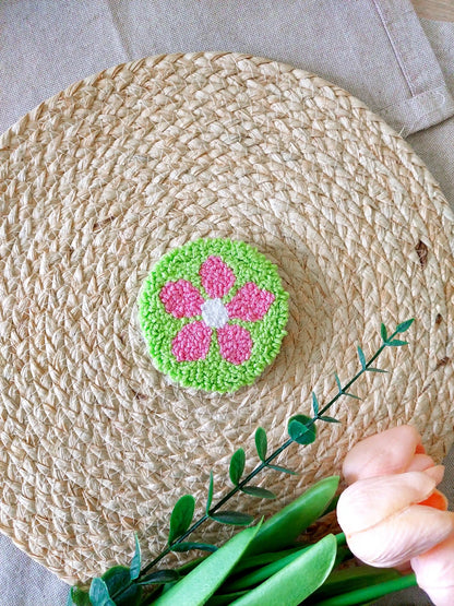 Handmade punch needle coaster featuring a bright green background with a pink flower, photographed on a woven mat with decorative flowers in the back.