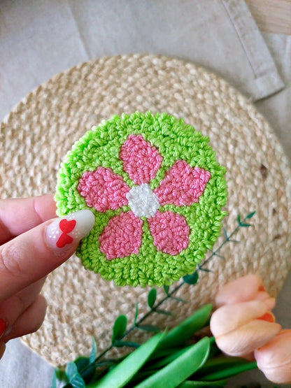 Handmade punch needle coaster featuring a bright green background with a pink flower, photographed on a woven mat with decorative flowers in the back.