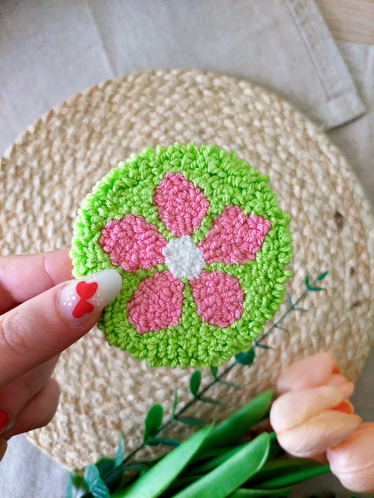 Handmade punch needle coaster featuring a bright green background with a pink flower, photographed on a woven mat with decorative flowers in the back.