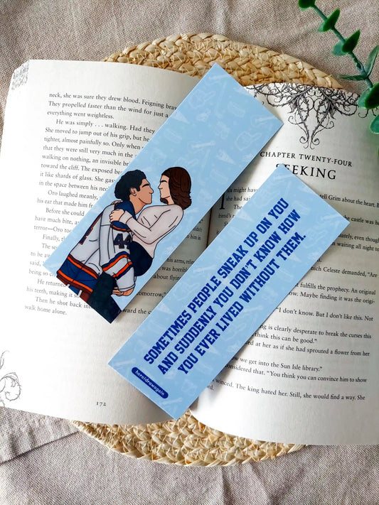 Off Campus inspired bookmark displayed on an open book, featuring an illustration of Graham and Hannah on the front and the quote "Sometimes people sneak up on you and suddenly you don't know how you ever lived without them" on the back.

