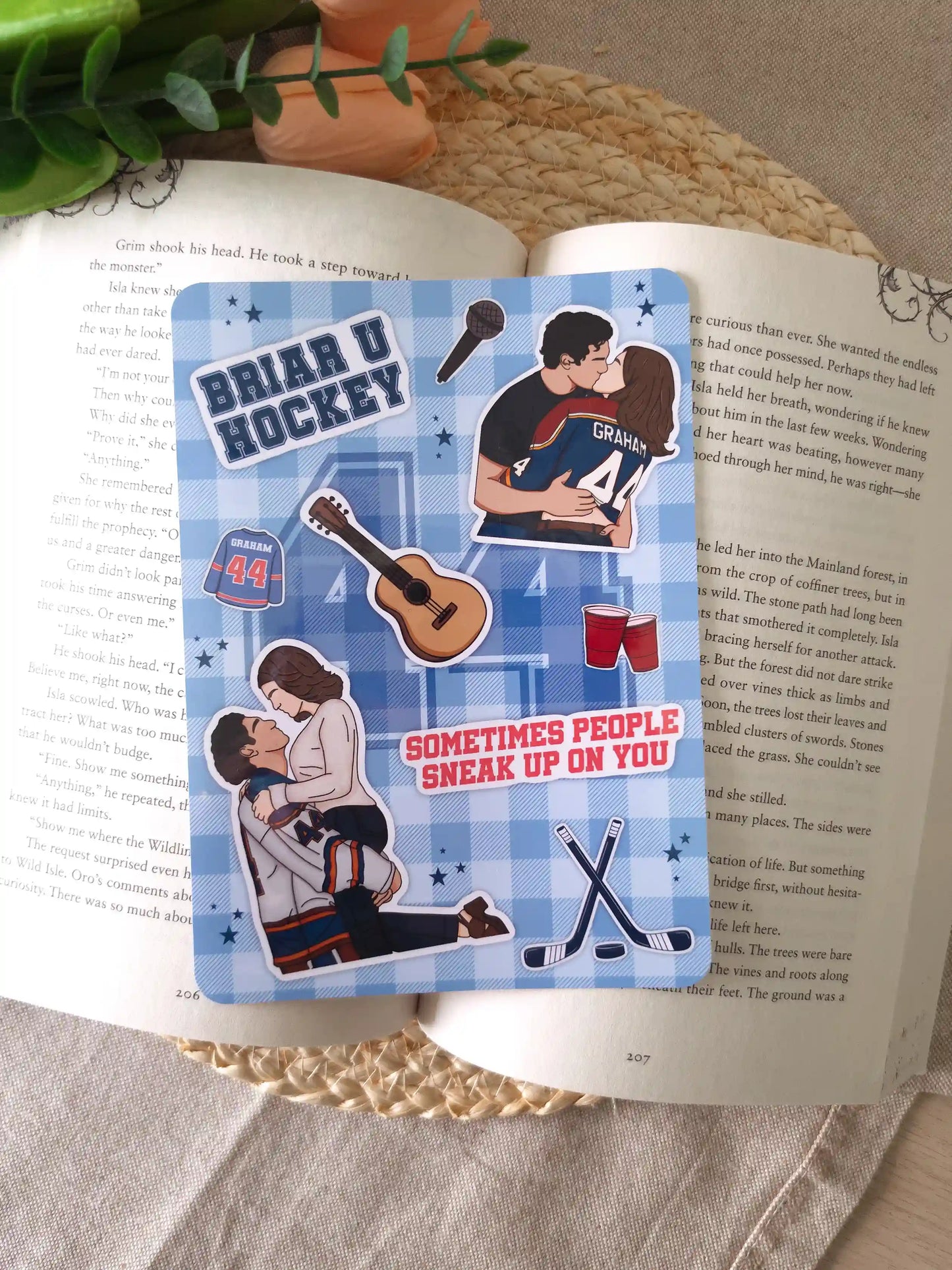 An e-reader insert with a blue gingham background featuring illustrated elements from The Deal by Elle Kennedy, including a "Briar U Hockey" badge, a Graham #44 jersey, two illustrations of Hannah and Garrett, crossed hockey sticks, a guitar, red party cups, a microphone, and the quote "Sometimes people sneak up on you" in red lettering, placed on an open book resting on a woven rattan mat. 