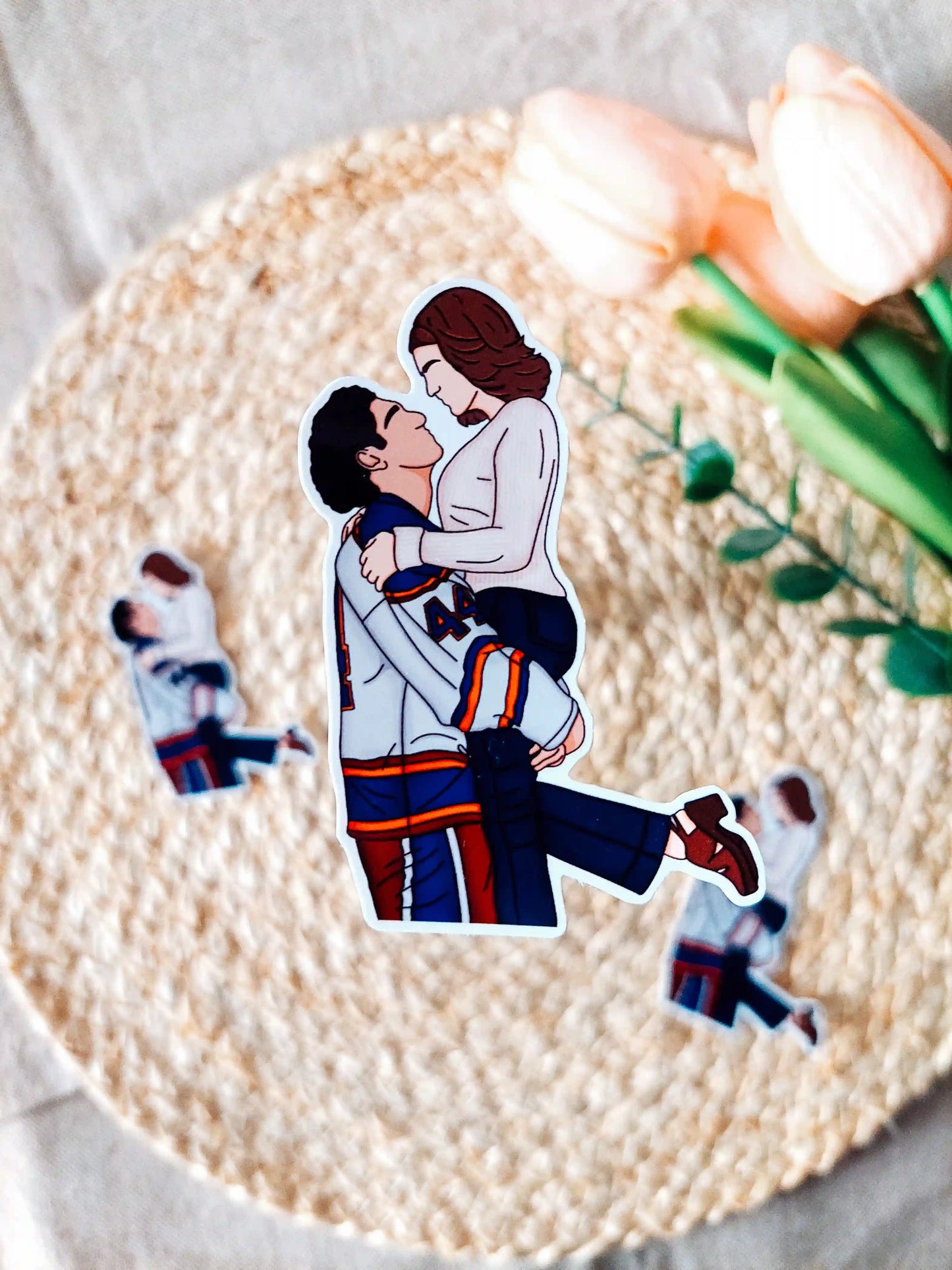 Off Campus inspired sticker photographed on a rattan placemat, featuring an illustration of Garrett in his hockey jersey lifting Hannah.