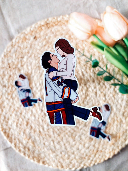 Off Campus inspired sticker photographed on a rattan placemat, featuring an illustration of Garrett in his hockey jersey lifting Hannah.