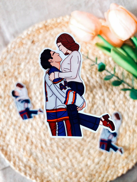 Off Campus inspired sticker photographed on a rattan placemat, featuring an illustration of Garrett in his hockey jersey lifting Hannah.