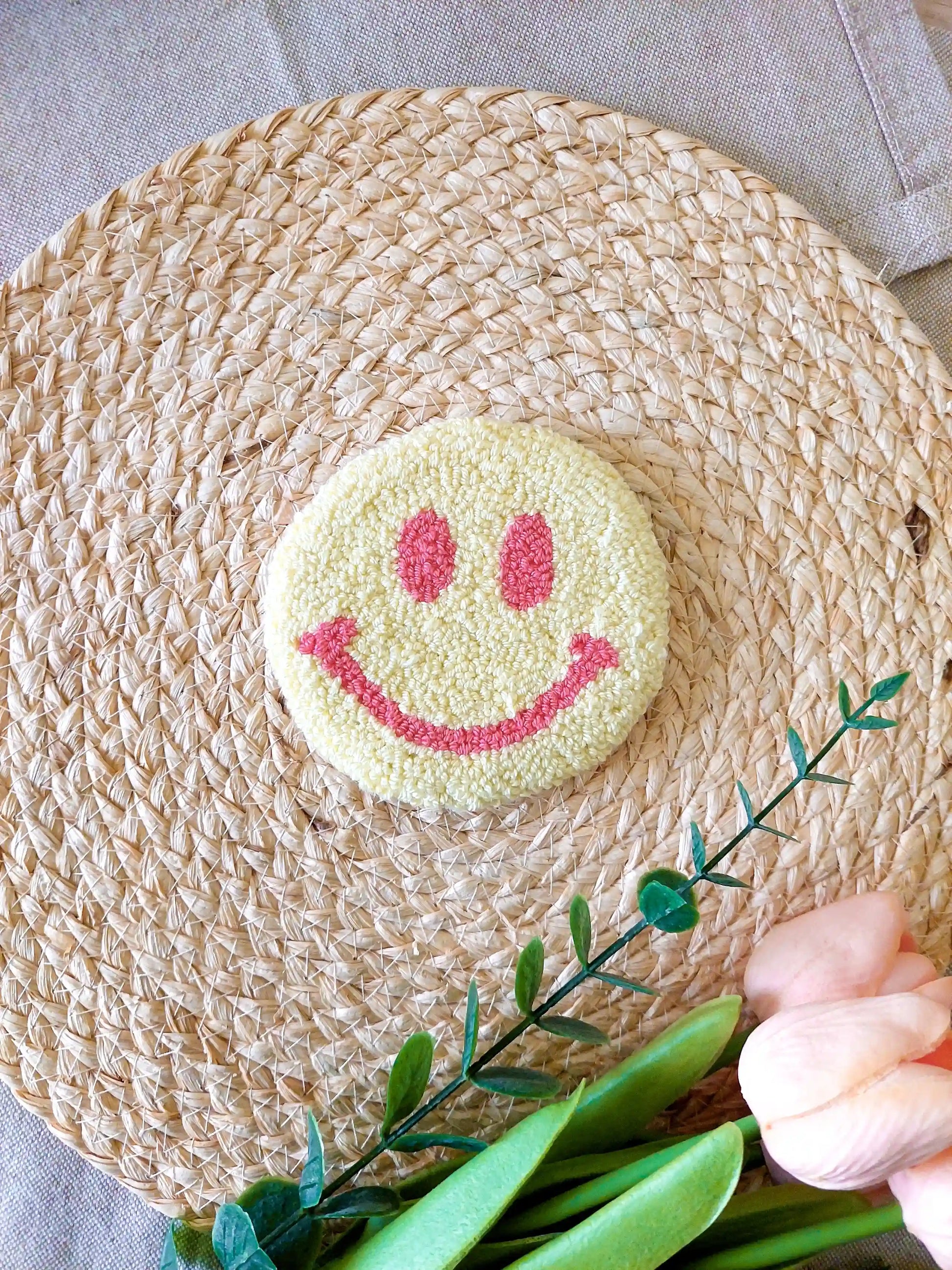 Handmade round punch needle coaster featuring a yellow and pink happy face design, photographed on a woven mat with decorative pink flowers in the back.