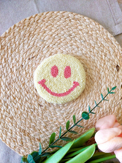 Handmade round punch needle coaster featuring a yellow and pink happy face design, photographed on a woven mat with decorative pink flowers in the back.