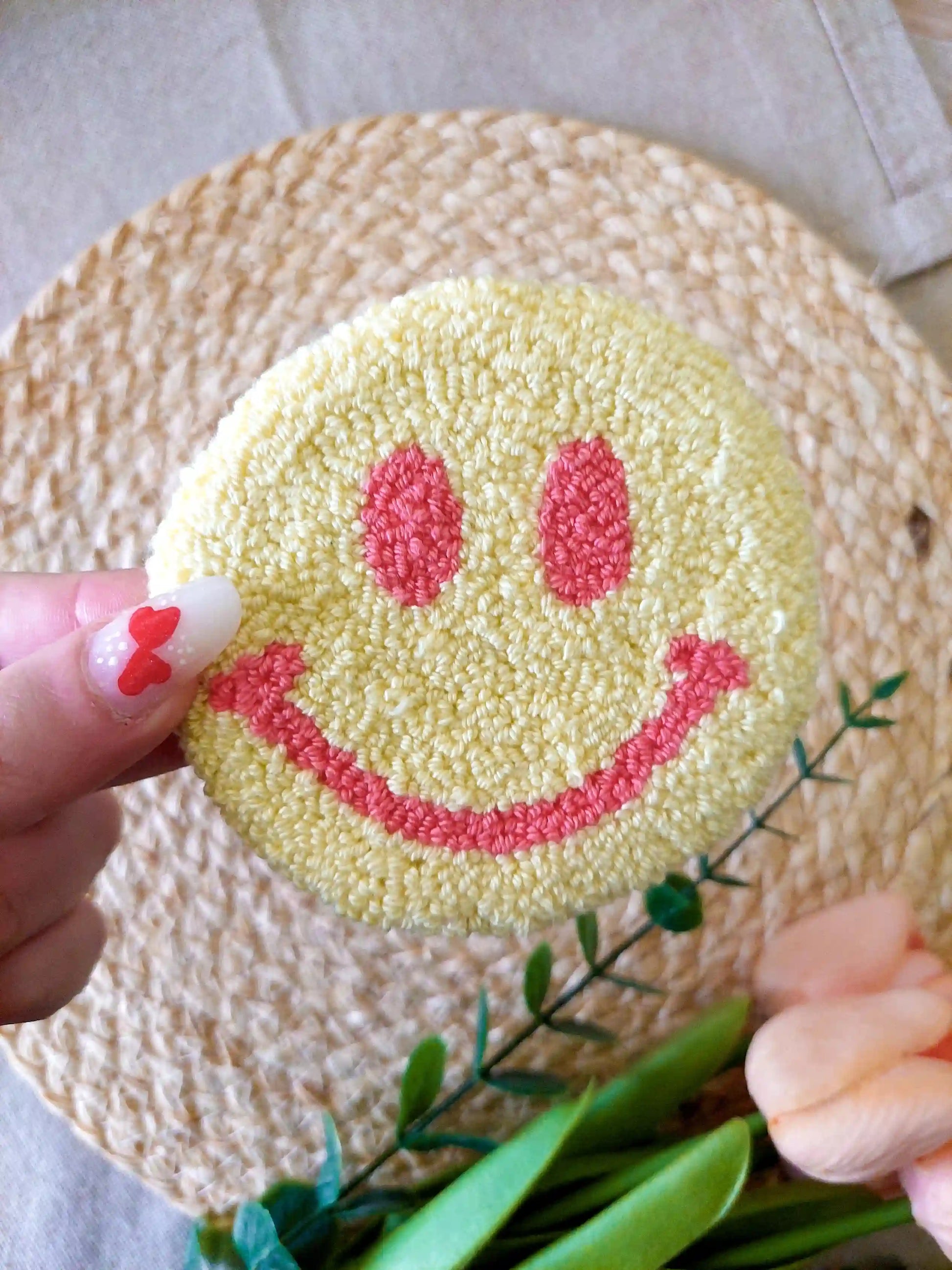 Handmade round punch needle coaster featuring a yellow and pink happy face design, photographed on a woven mat with decorative pink flowers in the back.