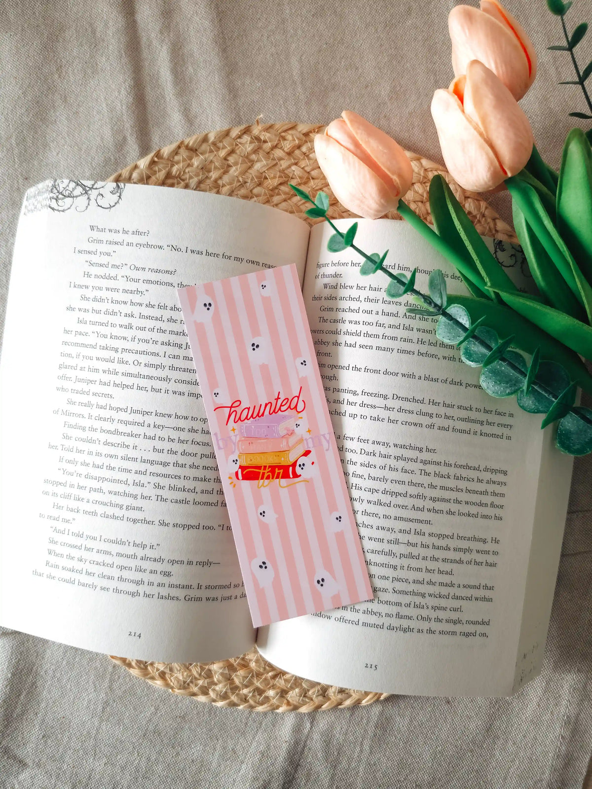 A laminated bookmark on a pale pink striped background featuring an illustration of a colorful stack of books with a small white ghost emerging from the top, the text "haunted by my tbr" cursive lettering, and small ghost face doodles scattered across the design, resting on an open book on a woven rattan mat, with peach tulips and green eucalyptus sprigs in the upper right corner.