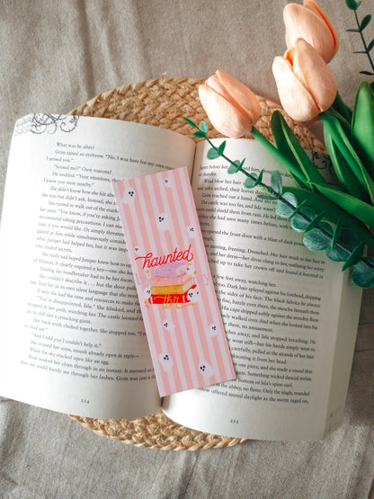 A laminated bookmark on a pale pink striped background featuring an illustration of a colorful stack of books with a small white ghost emerging from the top, the text "haunted by my tbr" cursive lettering, and small ghost face doodles scattered across the design, resting on an open book on a woven rattan mat, with peach tulips and green eucalyptus sprigs in the upper right corner.