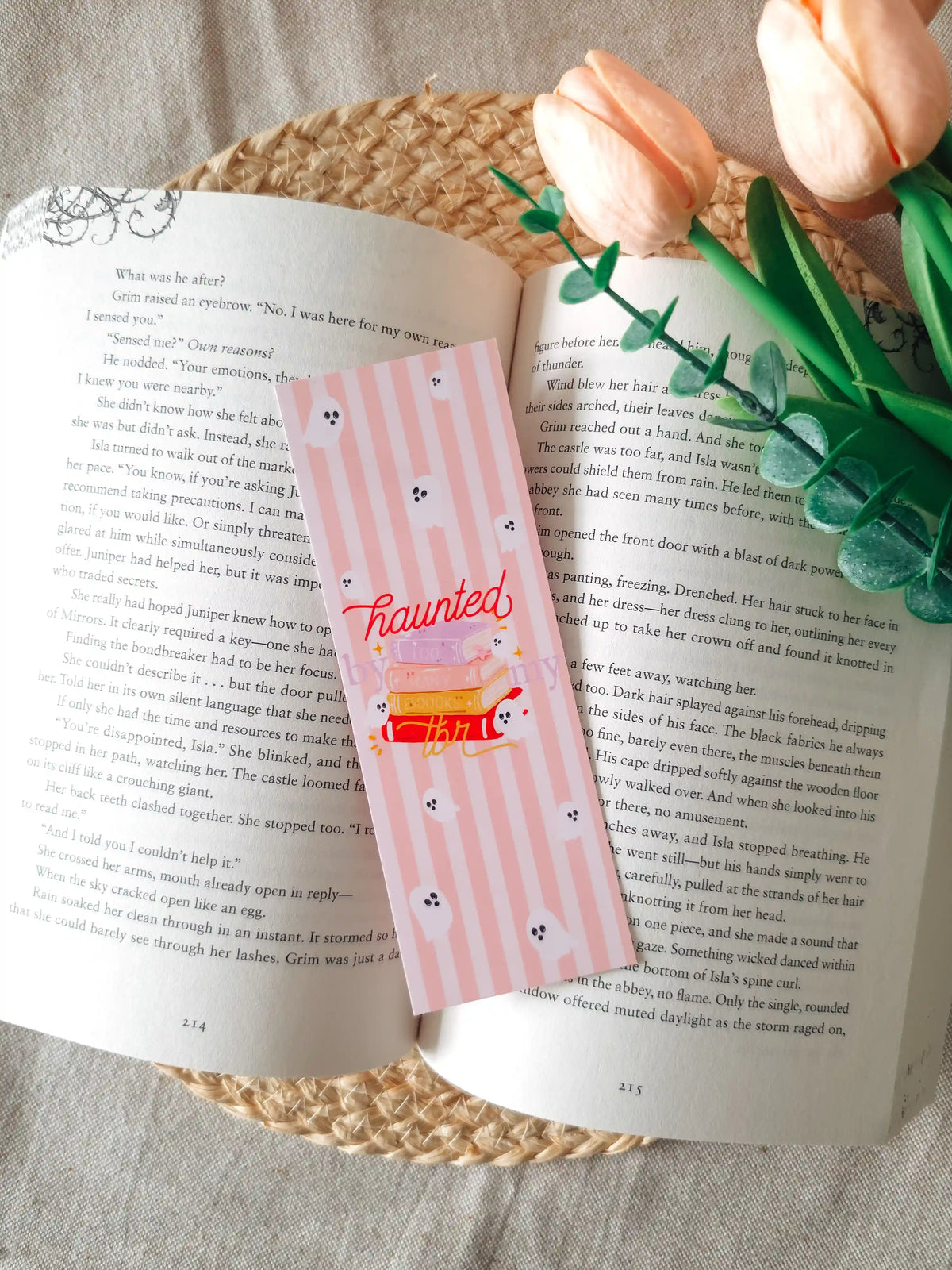 A laminated bookmark on a pale pink striped background featuring an illustration of a colorful stack of books with a small white ghost emerging from the top, the text "haunted by my tbr" cursive lettering, and small ghost face doodles scattered across the design, resting on an open book on a woven rattan mat, with peach tulips and green eucalyptus sprigs in the upper right corner.