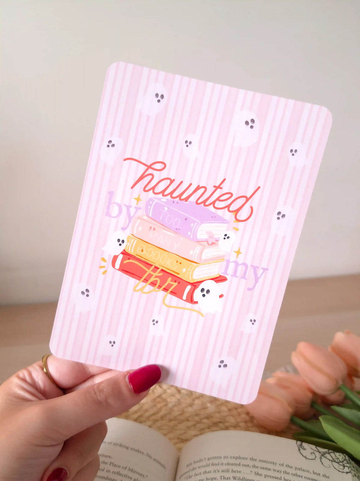 A laminated e-reader insert on a pale pink striped background featuring the text "haunted" in red cursive lettering at the top, an illustration of a colorful stack of books with a small ghost and gold sparkles, the text "by my TBR" in lighter and gold cursive script, and small ghost face doodles scattered across the background, resting on an open book on a woven rattan mat, with peach tulips and green leaves in the upper left corner.