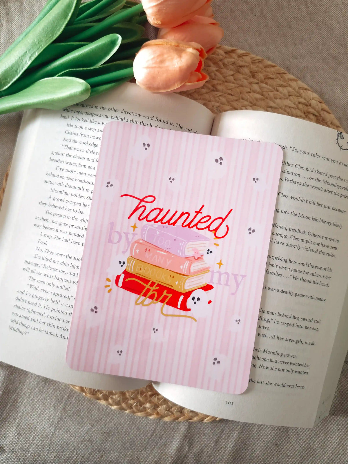 A laminated e-reader insert on a pale pink striped background featuring the text "haunted" in red cursive lettering at the top, an illustration of a colorful stack of books with a small ghost and gold sparkles, the text "by my TBR" in lighter and gold cursive script, and small ghost face doodles scattered across the background, resting on an open book on a woven rattan mat, with peach tulips and green leaves in the upper left corner.