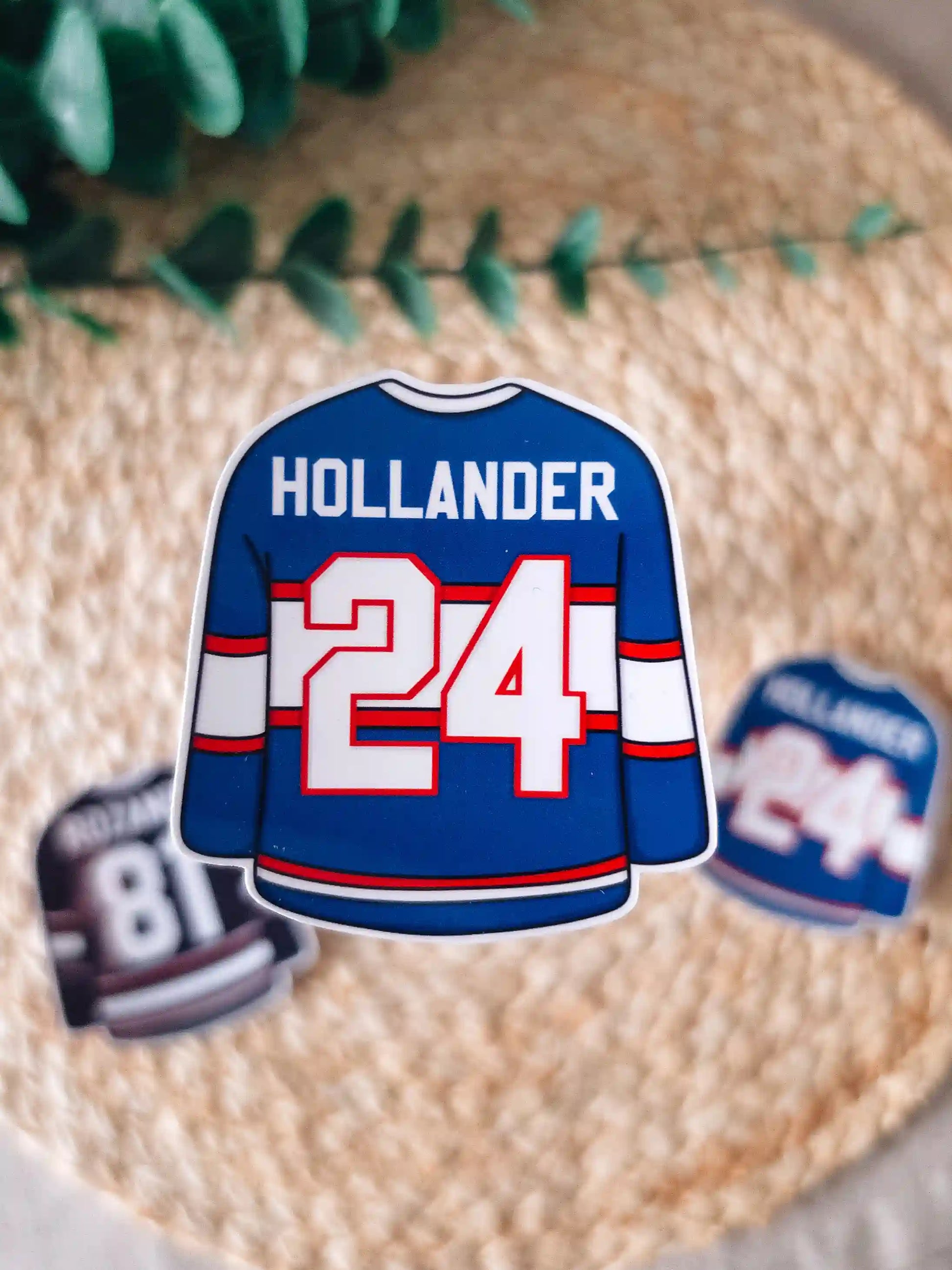 A die-cut sticker shaped like a blue hockey jersey reading "Hollander 24" in white lettering with red and white stripes at the center of a woven rattan mat. 