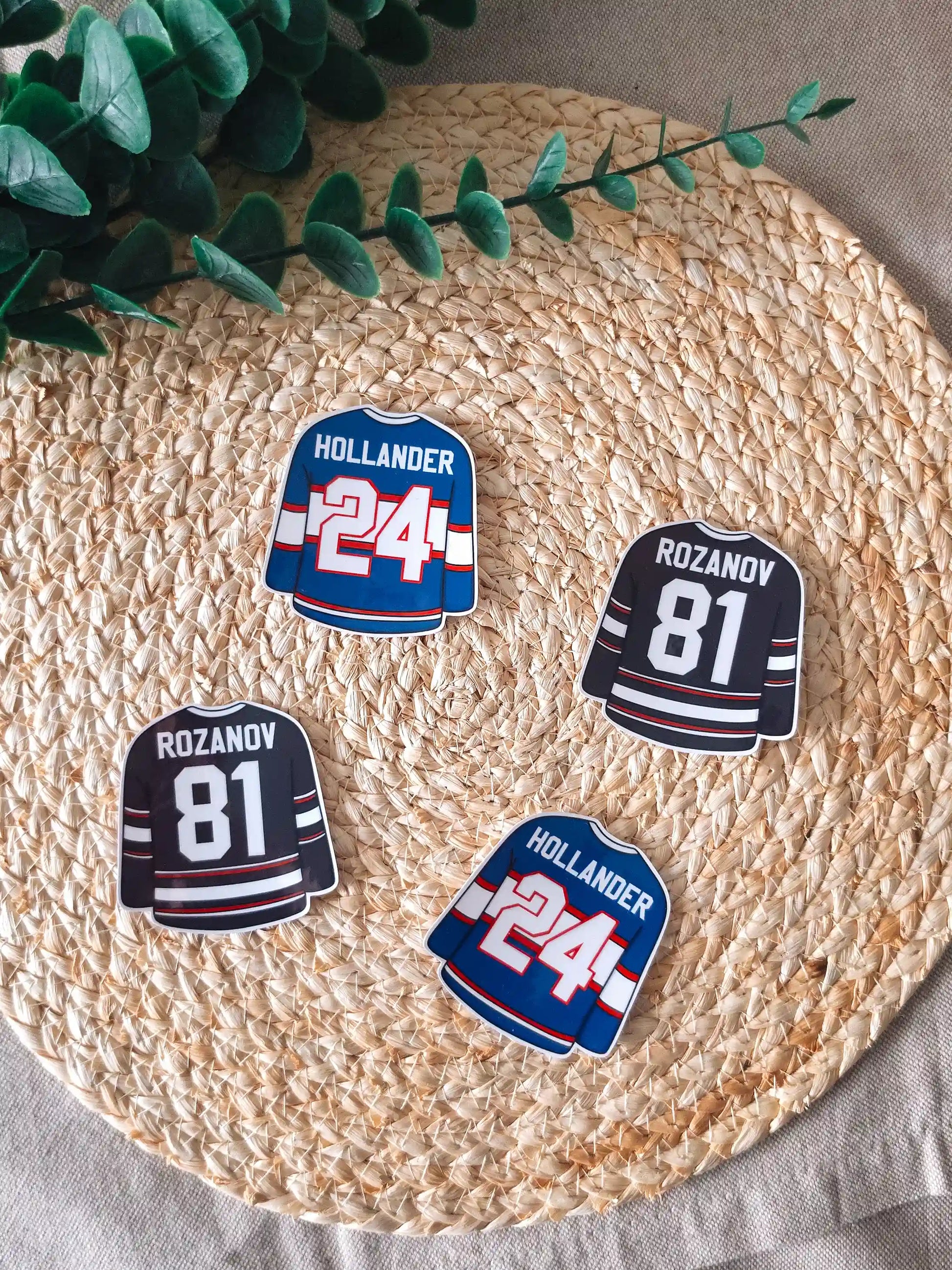 Four die-cut hockey jersey stickers arranged on a woven rattan mat, two black Rozanov #81 jerseys and two blue Hollander #24 jerseys, with green branches resting in the upper corner.
