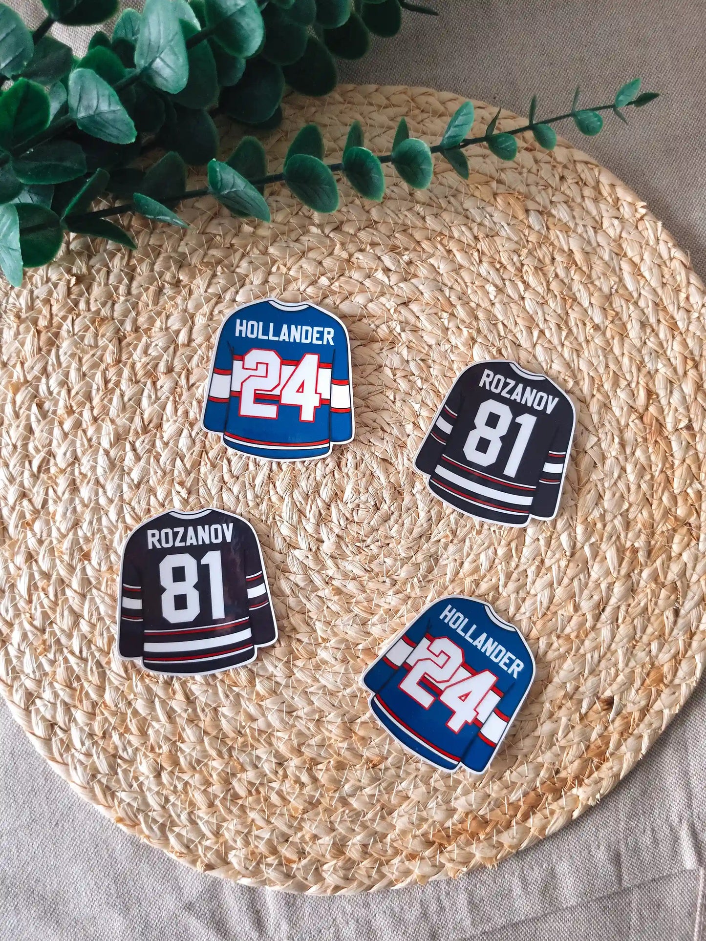 Four die-cut hockey jersey stickers arranged on a woven rattan mat, two black Rozanov #81 jerseys and two blue Hollander #24 jerseys, with green branches resting in the upper corner.