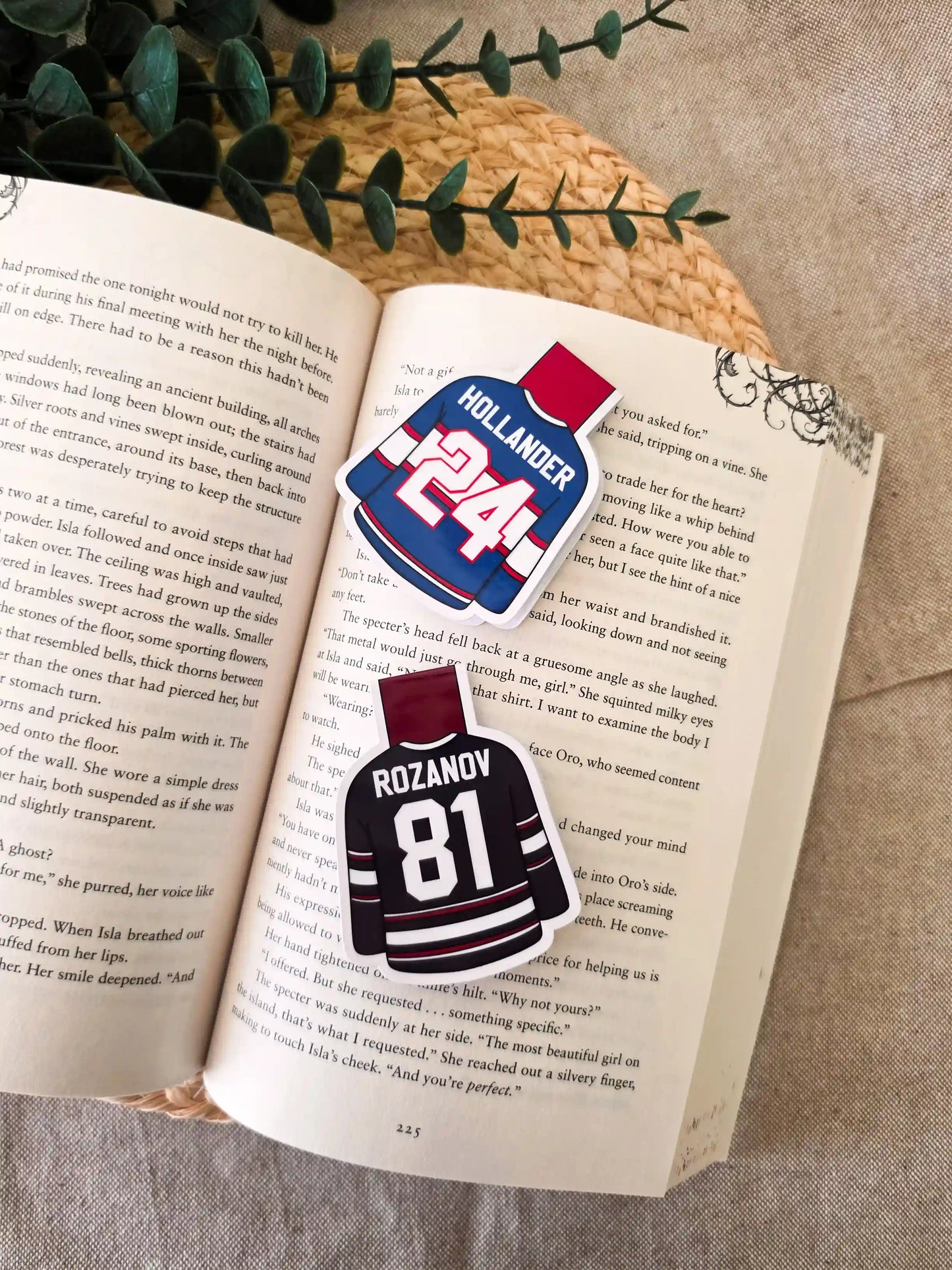 Two glossy laminated magnetic bookmarks shaped like hockey jerseys on an open book, one Hollander #24 blue Montreal jerseys and one Rozanov #81 black Boston jerseys from Heated Rivalry.