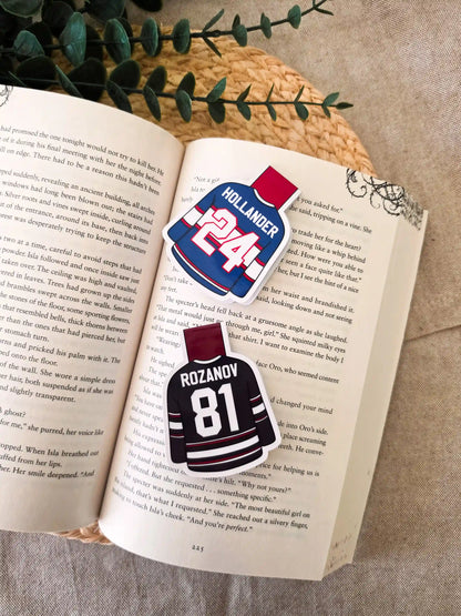 Two glossy laminated magnetic bookmarks shaped like hockey jerseys on an open book, one Hollander #24 blue Montreal jerseys and one Rozanov #81 black Boston jerseys from Heated Rivalry.