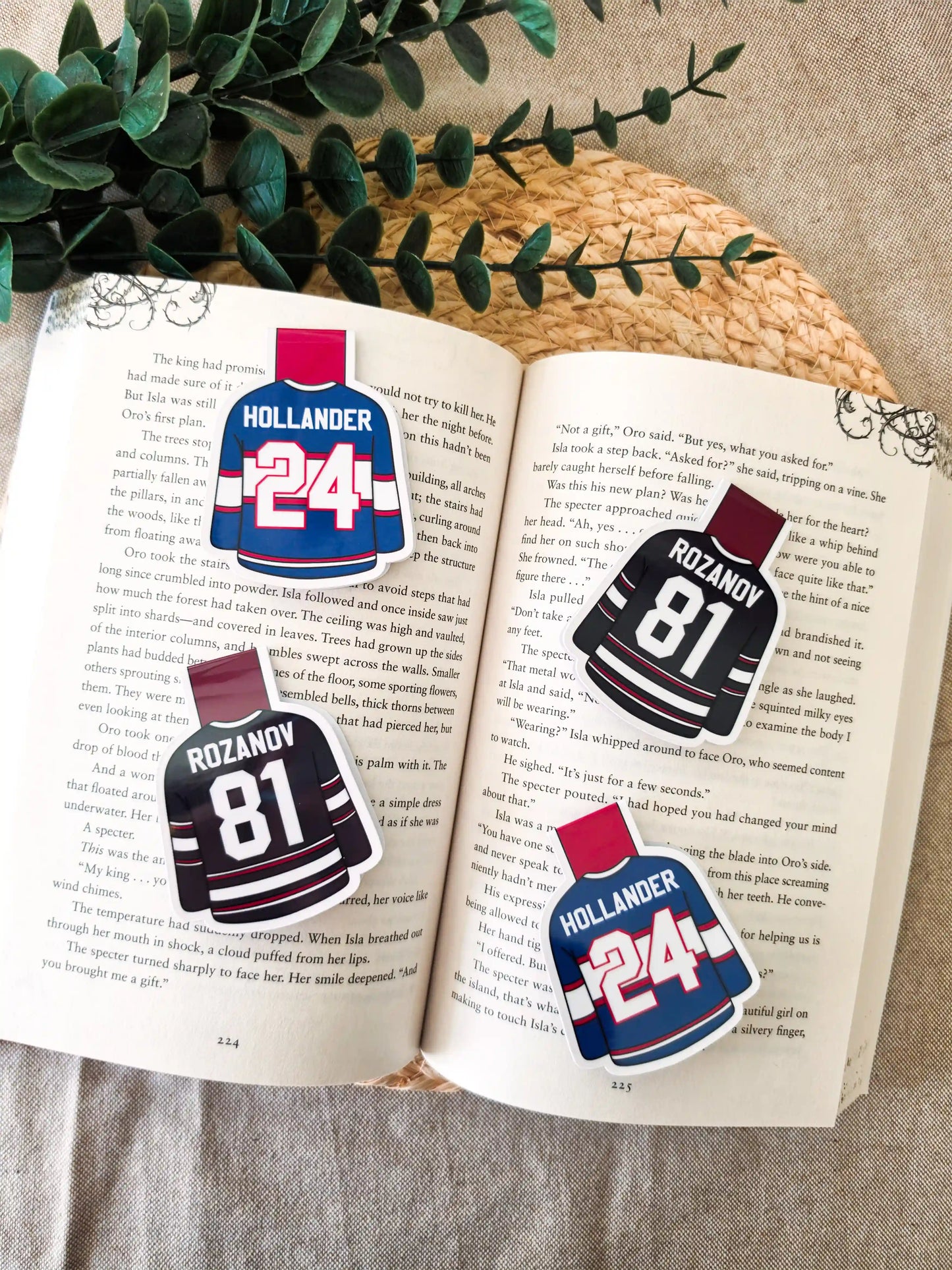 Four glossy laminated magnetic bookmarks shaped like hockey jerseys on an open book, two Hollander #24 blue Montreal jerseys and two Rozanov #81 black Boston jerseys from Heated Rivalry.