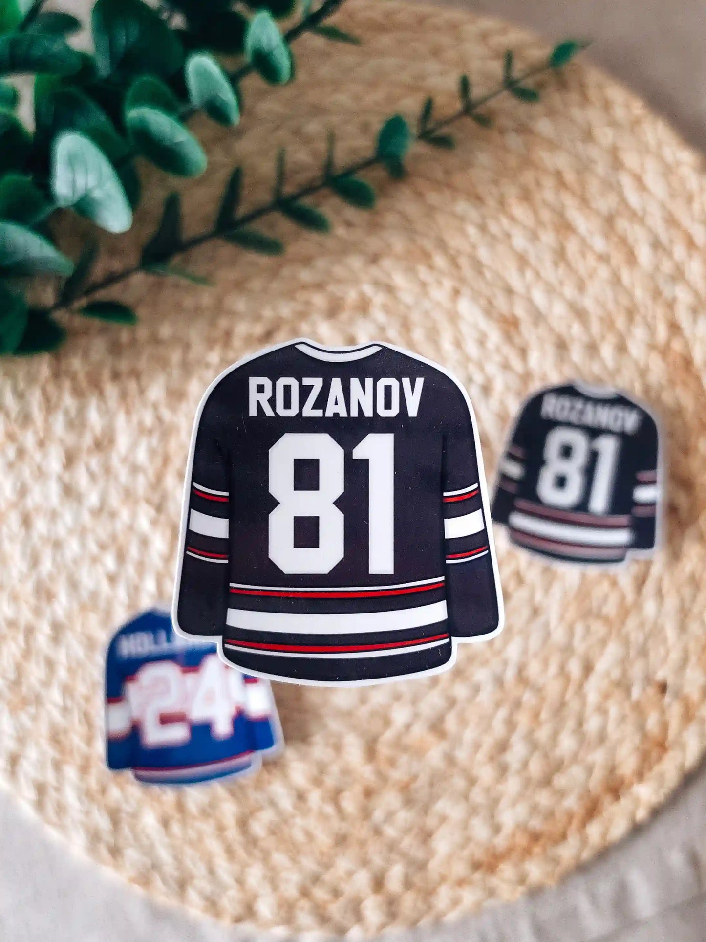 A die-cut sticker shaped like a blue hockey jersey reading "Rozanov 81" in white lettering with red and white stripes at the center of a woven rattan mat. 