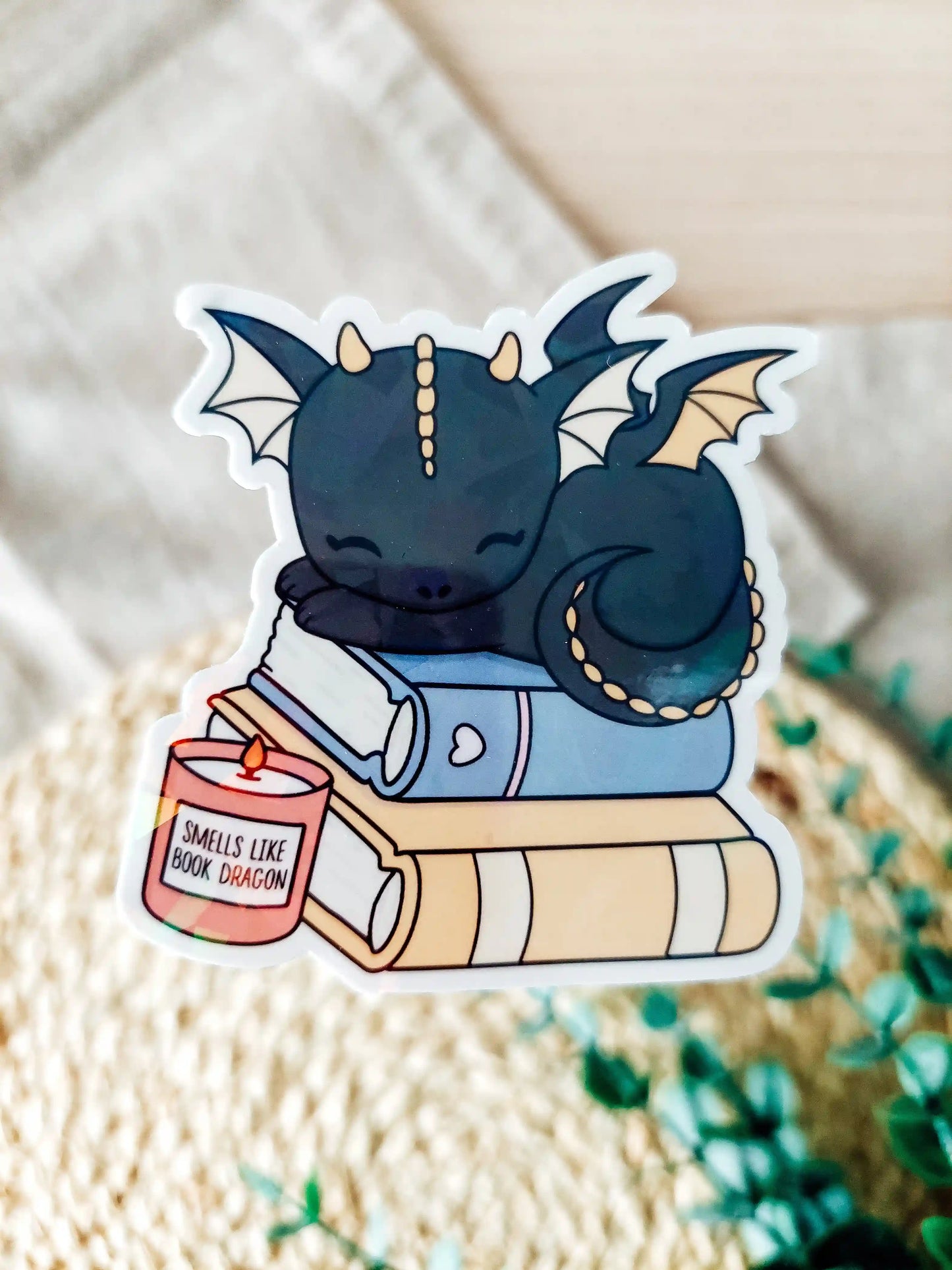Holographic vinyl sticker of a black baby dragon sleeping on stacked books with a candle.