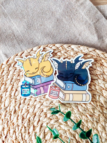 Two holographic vinyl stickers featuring sleeping baby dragons resting on stacked books with small candles, photographed on a woven neutral background.